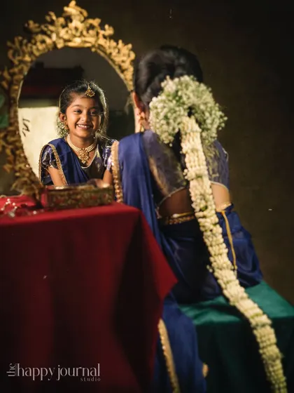 A girl in a traditional blue silk lehenga, with her hair braided with jasmine, smiles into a mirror. A beautiful reflection of joy and tradition.