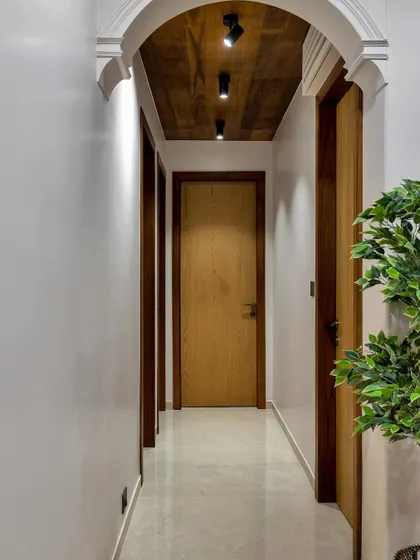 An arched hallway with a wooden ceiling and modern track lighting creates a dramatic yet warm transition between spaces. The clean white walls and simple wooden doors maintain a sense of minimalist elegance.