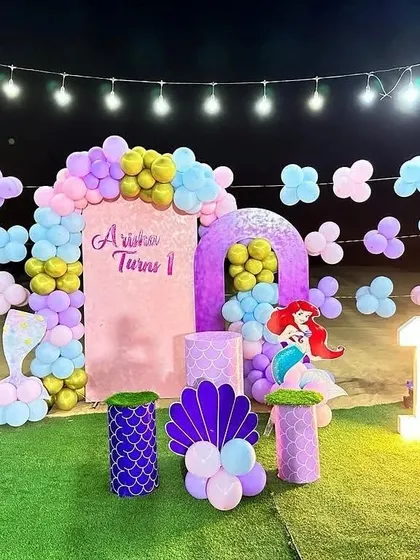 An outdoor 'Little Mermaid' first birthday party on an artificial grass lawn, featuring a marquee number '1' and themed props.