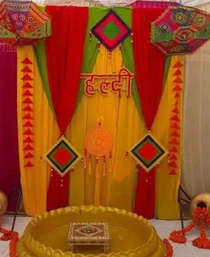 A Haldi backdrop with red, yellow, and green drapes, decorated with colorful kites and a central dreamcatcher.