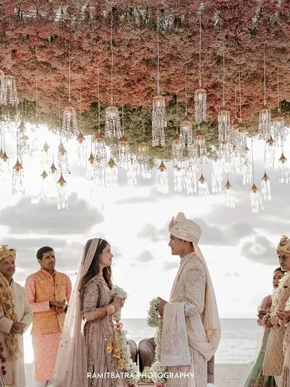 The couple stands under a stunning floral mandap for their sunset ceremony, with the ocean behind them.