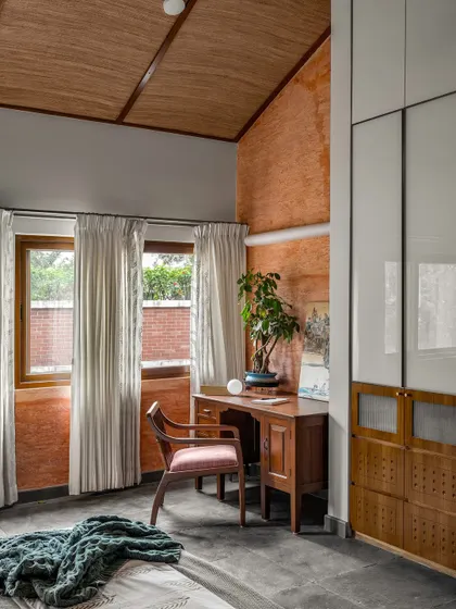This bedroom study corner combines rustic and modern elements, with an exposed terracotta wall, a vaulted ceiling, and sleek white wardrobes. It’s a warm, personal space designed for work and rest.