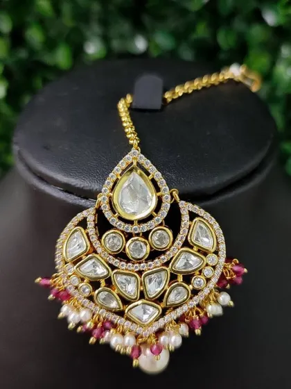 A classic kundan maang tikka with a teardrop-shaped central stone. The piece is finished with delicate pearl and red bead drops.