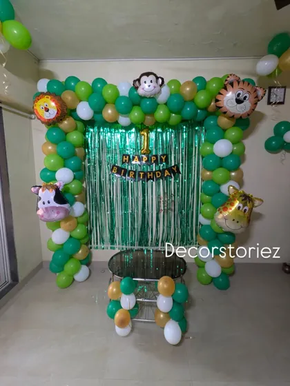A classic jungle theme balloon arch for a first birthday. This decoration uses green and gold balloons, a foil curtain backdrop, and friendly animal foil balloons like a monkey, lion, and giraffe.
