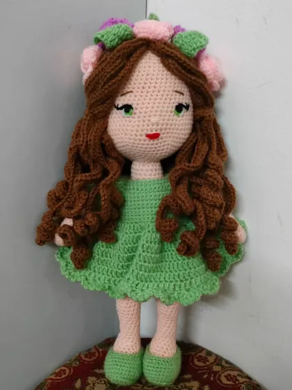 A beautiful doll with long, curly brown hair, a green dress, and a crown of pink and green flowers.