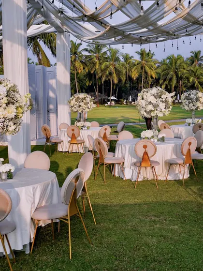 A beautiful daytime view of the reception setup on the lawns of the Taj Exotica, Goa. The crisp white tables are adorned with large floral centerpieces, creating an atmosphere of fresh, natural elegance against the backdrop of palm trees.