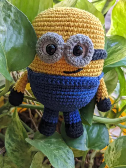 A cheerful Minion toy, crocheted with his classic blue overalls and big grey goggles, hiding among some green leaves.