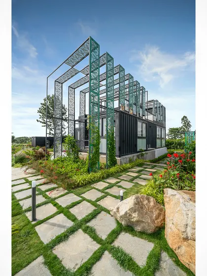 A daytime view of the container home, showing the green perforated metal structure that adds a sculptural element to the building. The surrounding greenery and stone pathway integrate the home into its landscape.