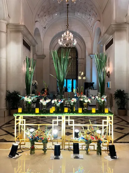 An elegant console table arrangement at The Leela Palace entrance. The golden table is decorated with an array of floral vases and candles, creating a luxurious welcome display.