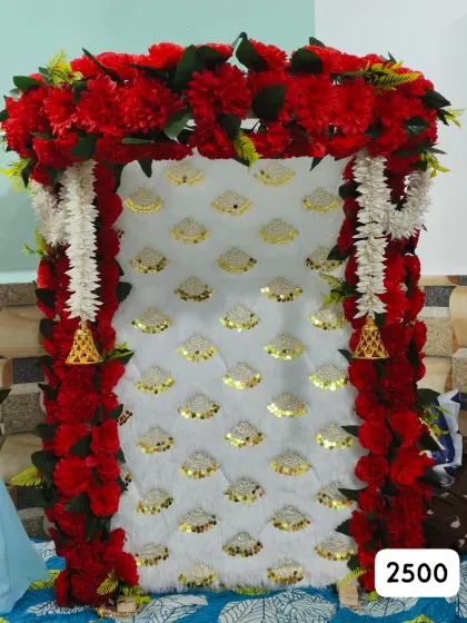 Our ₹2,500 ready-made Ganpati backdrop has red flowers around a white and gold panel, with white laris and bell accents.