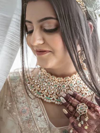 A serene bridal portrait. The bride looks down, showcasing her elaborate necklace and the delicate henna on her hands, capturing a moment of quiet contemplation.