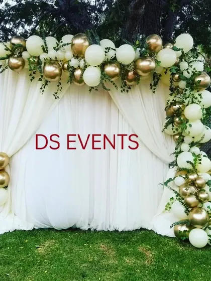 An elegant outdoor balloon decoration. This asymmetrical arch uses white and chrome gold balloons with fresh greenery tucked in, creating a sophisticated and modern photo backdrop for any celebration.