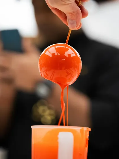 The art of the mirror glaze. A perfect, glossy orange sphere being dipped, a signature technique taught in many of our guest chef masterclasses.