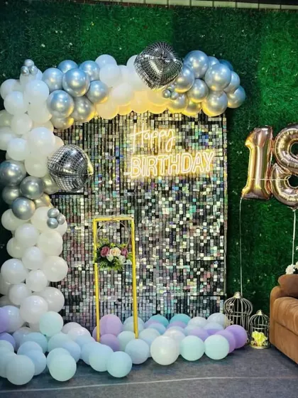 An 18th birthday party with a silver shimmer wall, a 'Happy Birthday' neon sign, and a balloon garland in silver and white.