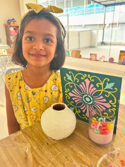 This young guest is all smiles with her collection of creations from a kids' art workshop, including a painted pot, a tile painting, and a cute froot loops candle.