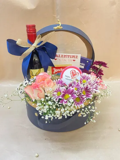 This stylish hamper in a navy blue container is perfect for him or her. It includes a bottle of wine and some treats, surrounded by a fresh arrangement of roses and daisies.