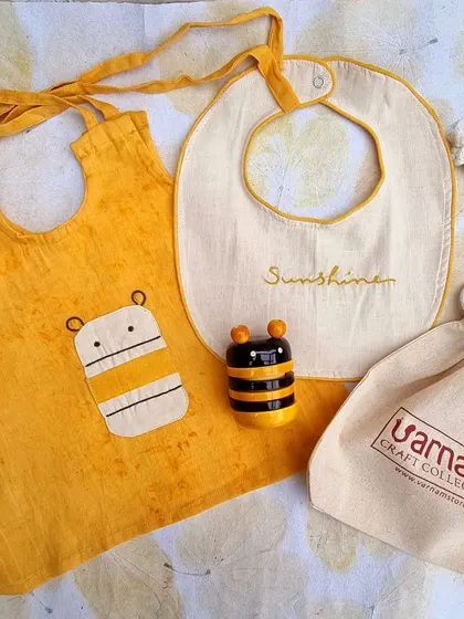 This newborn gift set features a bee-themed applique on a yellow hand-dyed Kora cotton dress, paired with a 'Bumbledore' bee rattle and a reusable kora pouch.