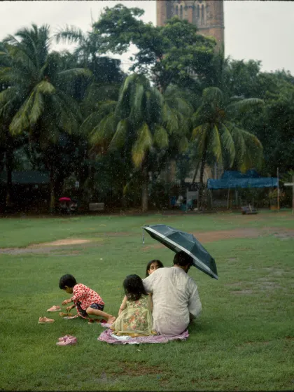 A family enjoys a picnic on the grass at Oval Maidan, huddled under an umbrella in the rain. It's a sweet, intimate moment of finding joy and making memories despite the weather.