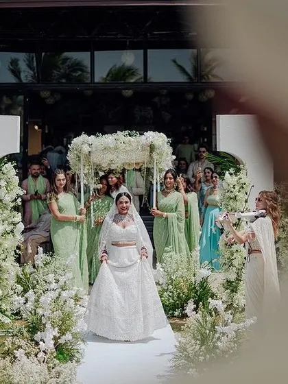 The bride walks down an aisle lined with white flowers, with her bridesmaids in green looking on.