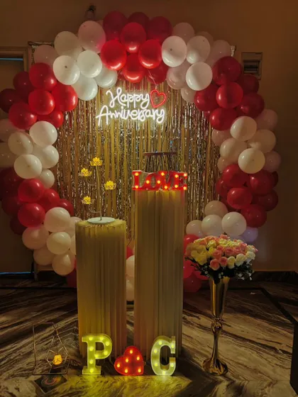 A classic anniversary decoration with a red and white balloon ring and a silver fringe backdrop. The 'Happy Anniversary' neon sign adds a warm glow to the celebration.