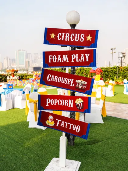A classic red and blue circus-themed signpost on a green lawn, directing guests to 'Foam Play', 'Carousel', and 'Popcorn'.