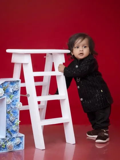 A classic first birthday photo with a bold red backdrop. The contrast between the red background, the white ladder, and the baby's dark outfit makes this a visually impactful shot.