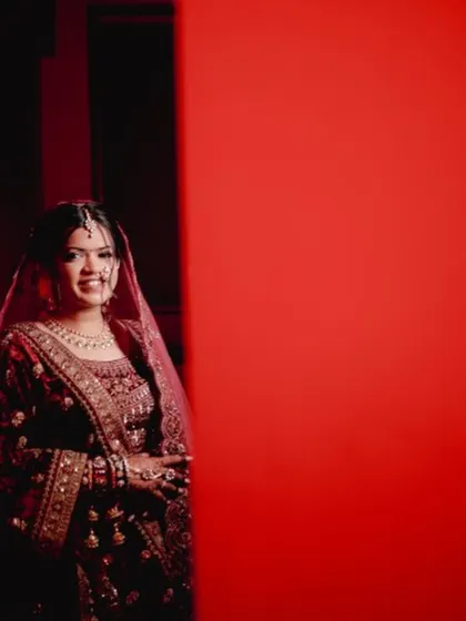 A creative shot using a bold red background to frame the bride. Her traditional maroon velvet lehenga and soft, glowing makeup create a striking contrast.