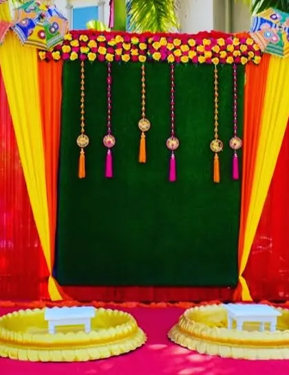 An outdoor Haldi setup with two 'urli' stations against a backdrop of red and green drapes, decorated with hanging tassels and umbrellas.