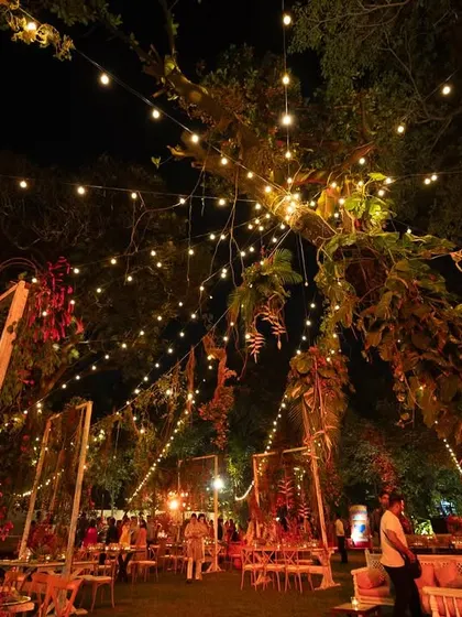An evening view of the Lohri celebration, where strings of fairy lights create a magical canopy over the garden. The warm, rustic setup with wooden tables and chairs is made festive with red-hued floral and vegetable decor.