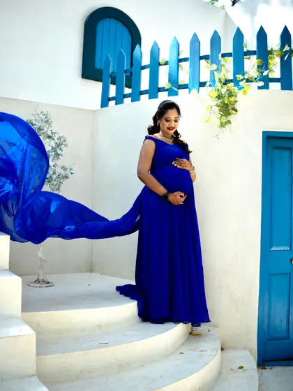 A dramatic shot of a mother-to-be in a flowing royal blue gown, with the fabric catching the wind against a Santorini-inspired white and blue backdrop.