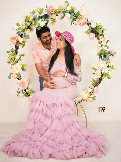A sweet couple's portrait in front of our floral hoop. The mom-to-be is seated, wearing a pink lace gown and hat, while her partner stands lovingly beside her.