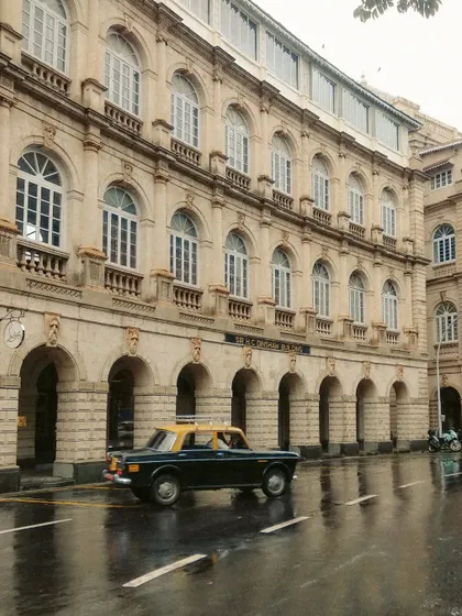 A kaali-peeli taxi on a wet street in front of a colonial-era building in Fort. The reflection on the road and the classic architecture create a scene rich with history.