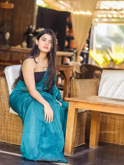 A modern take on saree styling. The model wears a teal saree paired with a contemporary black top, shot in a cafe with earthy tones. This shows how traditional wear can be styled for a modern, chic look.