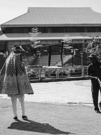 An unusual and artistic behind-the-scenes shot. The bride's lehenga is being prepared while a worker cleans the grounds, creating a juxtaposition of glamour and everyday life, made more poignant in black and white.