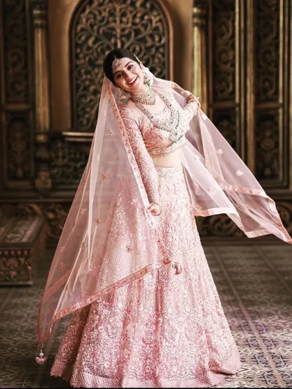 A full-length shot of Megha Shetty in the breathtaking pink bridal lehenga. The flowing veil and the sheer volume of the embroidered skirt create a truly unforgettable bridal silhouette.