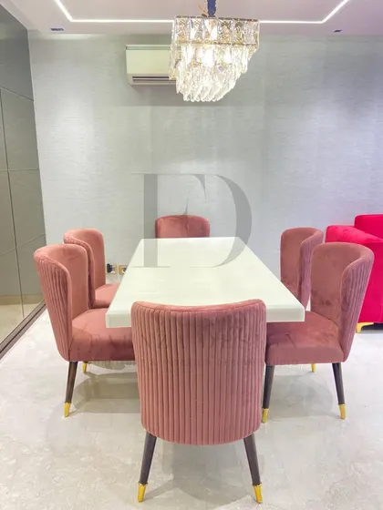 A front view of the dining set, highlighting the beautiful contrast between the pink chairs and the bright pink sofa in the background. The crystal chandelier adds sparkle and sophistication.