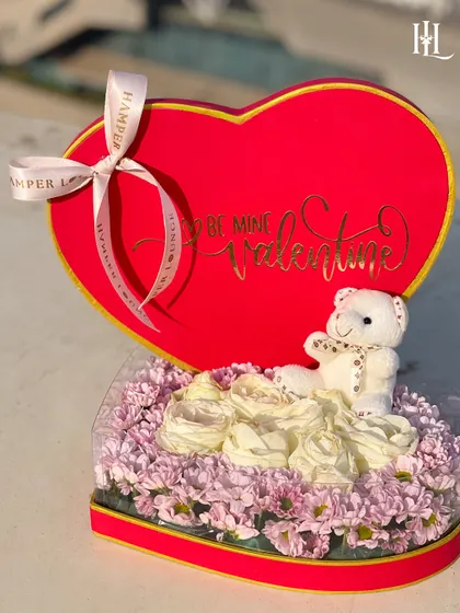 A heart-shaped floral hamper for your Valentine. This box is filled with soft white and purple flowers and a cuddly teddy bear, with "Be Mine Valentine" written in gold script.