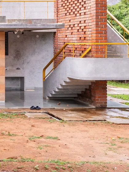 The cantilevered concrete staircase at "Aamra" appears to float alongside the exposed brick jaali wall. The yellow handrail adds a vibrant accent to the earthy tones of the structure.
