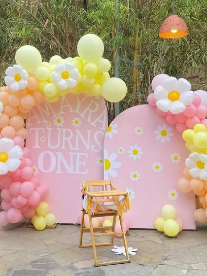 A sweet and cheerful Daisy Flower theme for a first birthday. This outdoor setup uses pink, yellow, and peach balloons with large daisy flower accents and a light-up number '1'.