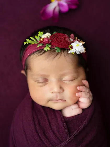 A close-up portrait focusing on the baby's serene expression and the beautiful floral headband that complements the deep plum wrap.