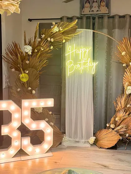 A boho-chic 35th birthday at home. This decor features a circular arch decorated with dried palm leaves and white florals, a "Happy Birthday" neon sign, and large marquee numbers.