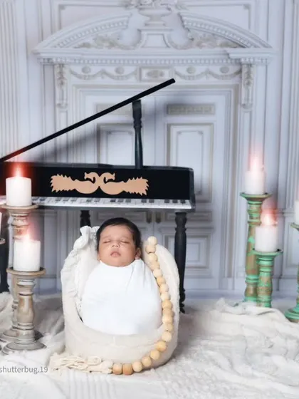 A swaddled newborn rests in a basket in front of the piano. The warm glow from the candles creates a serene and classical atmosphere.