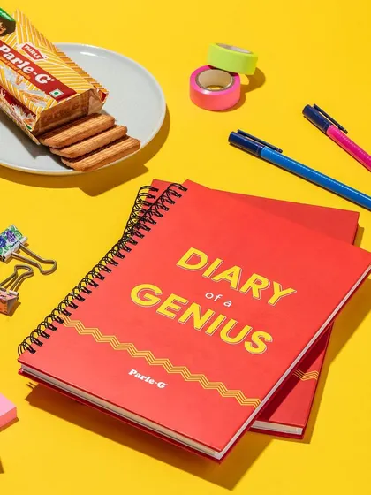 This flat lay shot for the Parle-G "Diary of a Genius" notebook uses bright, complementary colors and relevant props. The composition is playful and organized, perfectly matching the brand's fun aesthetic.