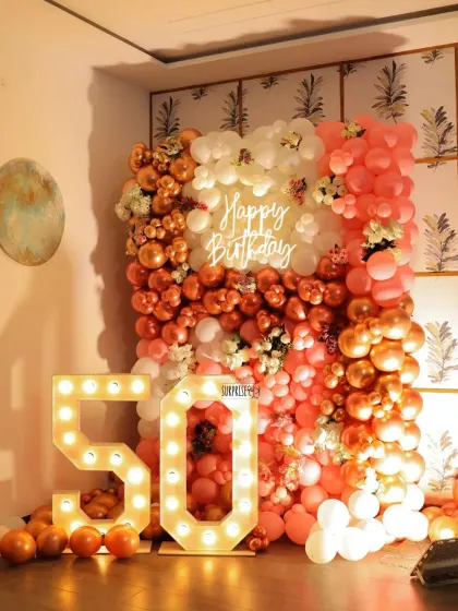 A beautiful setup for a 50th birthday celebration. This decor features a full wall of rose gold, pink, and white balloons, a "Happy Birthday" neon sign, and large marquee numbers, creating a stunning photo corner.