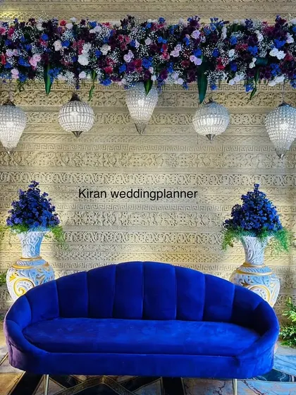 A chic and modern engagement or sangeet stage with a royal blue sofa, set against a textured gold wall and accented with blue and purple floral arrangements.