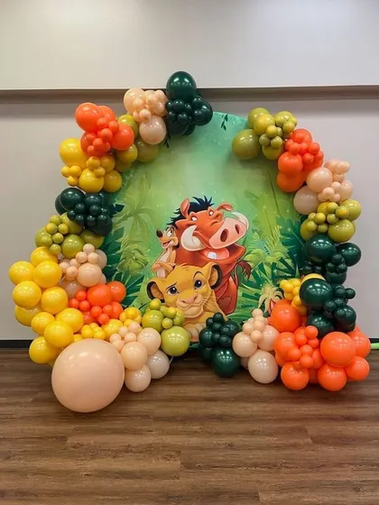 A simple yet effective Lion King themed backdrop. A circular panel with Simba, Timon, and Pumbaa is framed by an organic balloon garland in jungle-inspired colors.