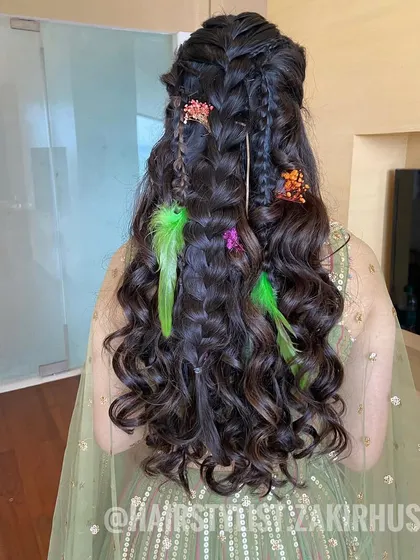 This bohemian hairstyle features multiple small braids, loose curls, and is decorated with colorful floral clips and bright green feathers. It's a fun and festive look for a Mehendi party.