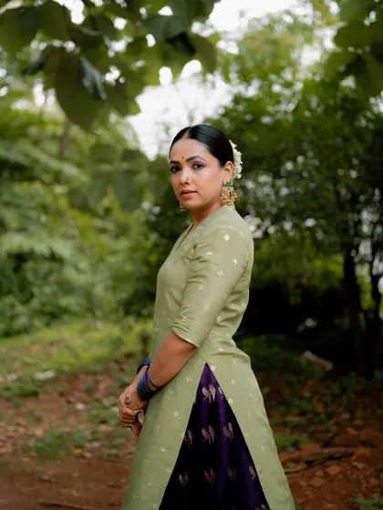 A full-length portrait of actress Anupama Gowda in a traditional green and purple outfit. The outdoor, natural setting complements the earthy tones of her attire.