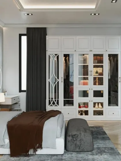 A bright and airy bedroom featuring a large, white wardrobe with elegant glass-panelled doors and a matching vanity table.