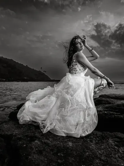A dramatic black and white bridal portrait by the sea. The moody sky and the texture of the rocks contrast beautifully with the soft, flowing wedding dress, creating a powerful and timeless image.